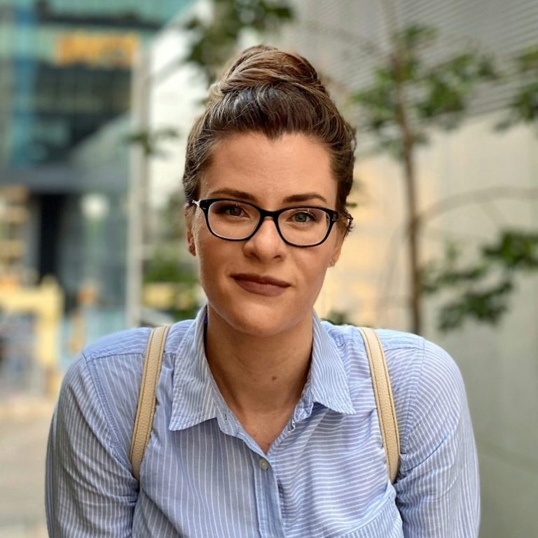 Young woman with glasses and a bun, wearing a blue shirt, smiling outdoors.